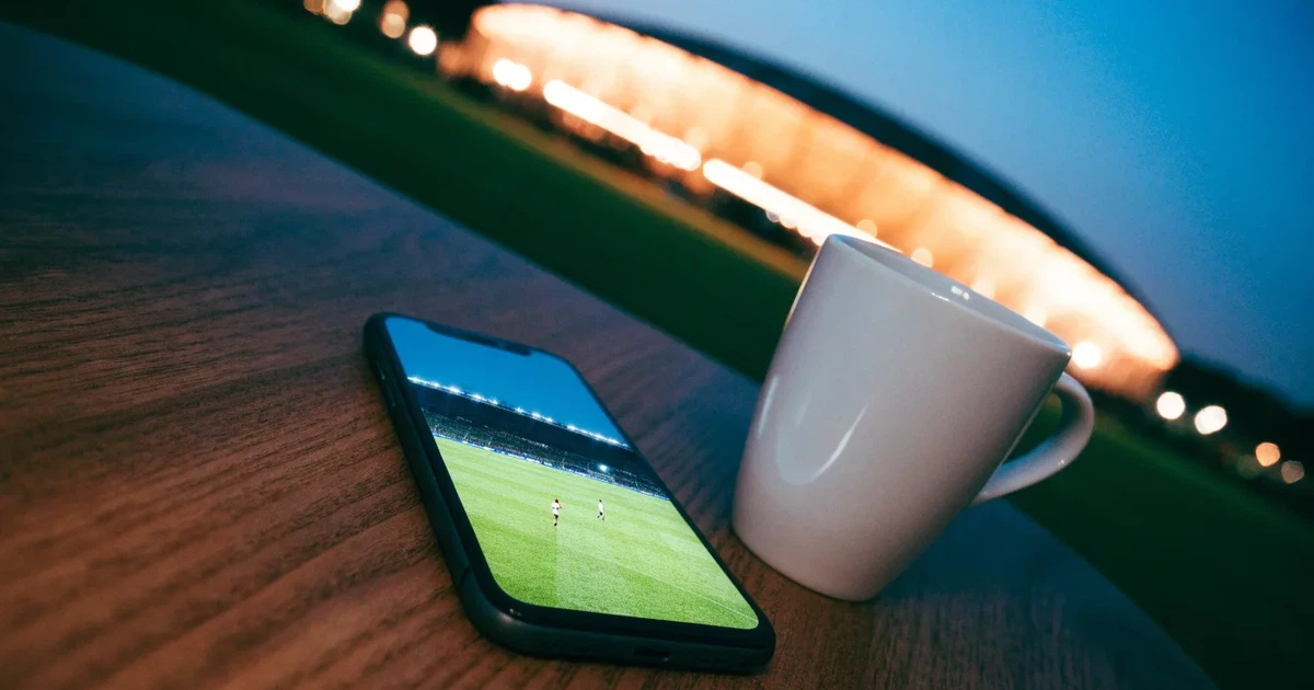 Ein Fußballstadion unter Flutlicht aus großer Entfernung gesehen, die Lichtkegel stehen im dunklen Abendhimmel über leeren Parkplätzen.